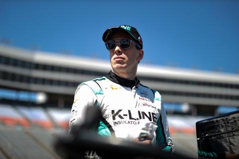 Dalton Kellett standing on the track in his racing outfit and baseball cap, wearing sunglasses.