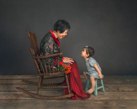 Asian woman, sitting in a rocking chair, looking down at a toddler, sitting on a blue stool.