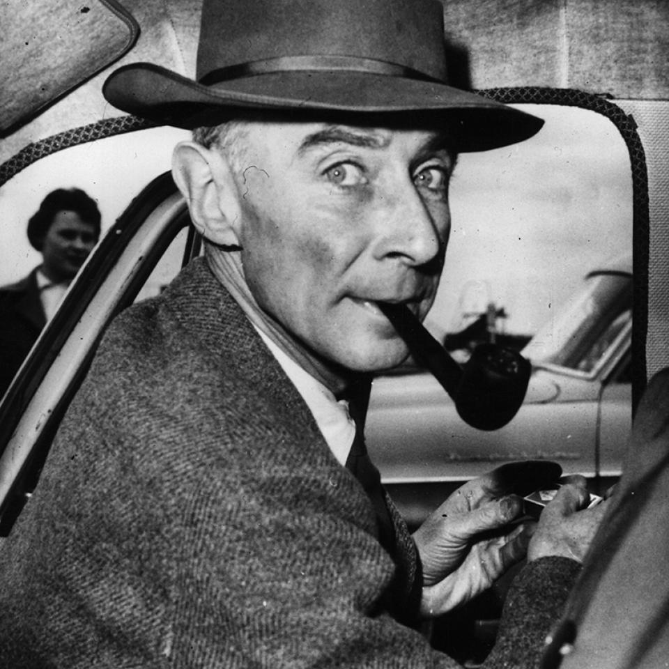 Robert Oppenheimer sits inside a car wearing a hat and smoking a pipe.
