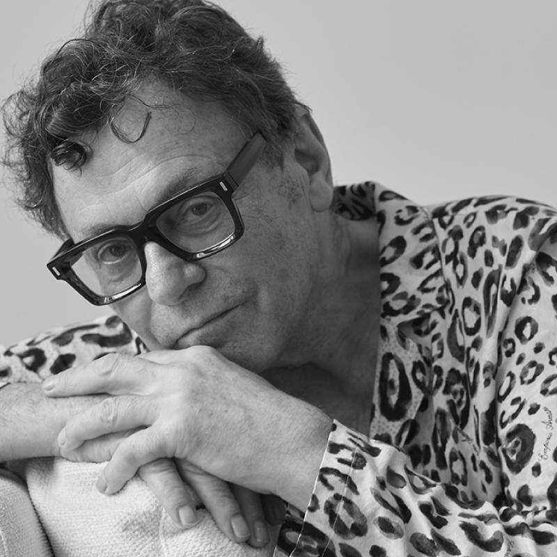 Man wearing thick-rimmed glasses and leopard pajamas, rests with his hands overlapping each on the back of a chair. His chin rests on his hands.