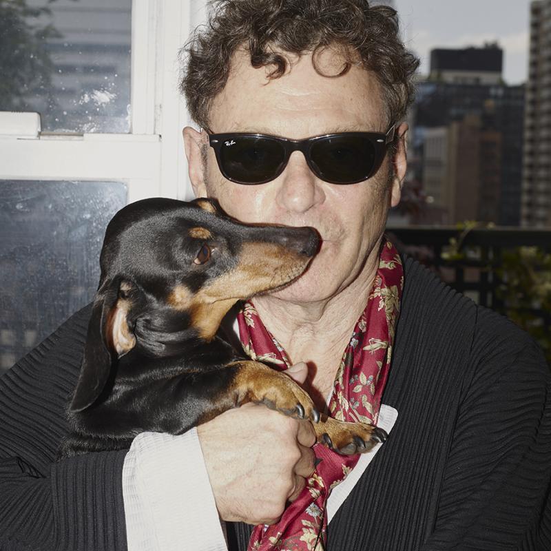 Man stands in front of windows with city buildings in the background. He is wearing dark sunglasses, a scarf around his neck, and a suit. He is holding a small dachshund up to his face..