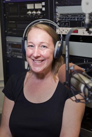 A person smiling with headphones on sitting in front of a microphone