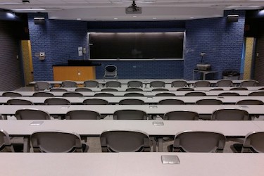 Classroom with rows of grey tables and swing chairs. The front is painted blue and has a large blackboard.