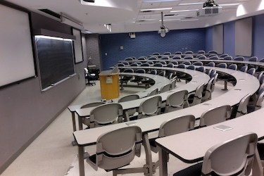Side view of a classroom. Grey tables and chairs in an arc shape around the podium and blackboard at the front.
