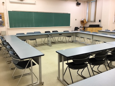 Classroom with tables and chairs arranged in a square. A podium is in the corner of the room and a large blackboard along the front wall.
