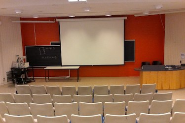 View of an auditorium from the back of the room. At the front is a white screen lowered in front of an orange wall.
