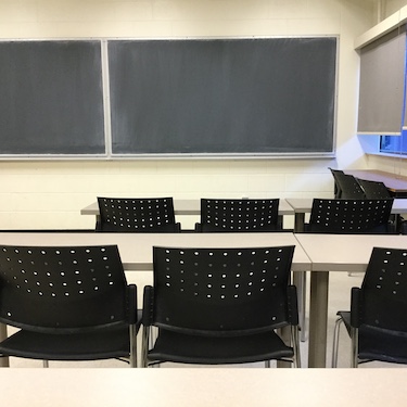 Narrow moveable tables with standard black moveable chairs set up in rows facing a chalkboard