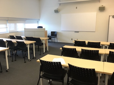 Classroom with narrow tables and black chairs. A large whiteboard and instructor podium at the front of the room.