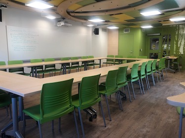 View from the side of the room: Narrow moveable tables with standard  moveable chairs set up in a rectangle with a whiteboard on one wall.