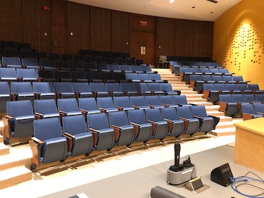View of a large classroom from the front of the room: Rows of fixed chairs with tablet tables attached. The walls are a bright yellow..