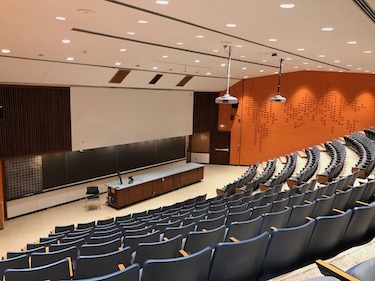 View of the front of the room: Rows of fixed chairs with tablet tables attached and screen at the front. The walls are a bright orange..