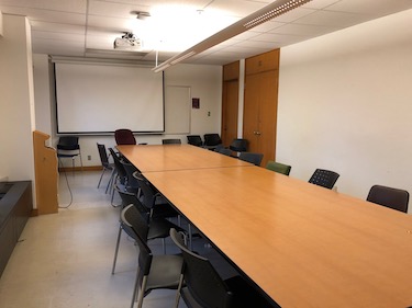 View from the side of the room: 2 large tables in the center of the room with moveable chairs surrounding them with a projector screen at the end of the room.