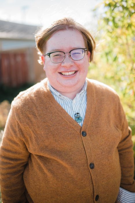 a photo of Drumlin from the waist up. They are a white nonbinary person with short light brown hair. They are wearing glasses, a brown cardigan sweater, and a white and green striped shirt. They have a necklace on with a turquoise pendant. They are smiling at the camera. 