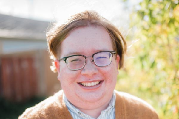 a photo of Drumlin from the waist up. They are a white nonbinary person with short light brown hair. They are wearing glasses, a brown cardigan sweater, and a white and green striped shirt. They have a necklace on with a turquoise pendant. They are smiling at the camera. 