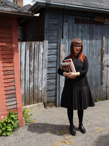 An image of a woman with red hair wearing a black dress and glasses holding books standing in front of an old wooden building