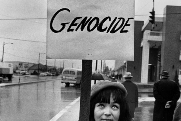 An image of a young Indigenous woman standing on a street with a sign that reads "Stop Cultural Genocide"