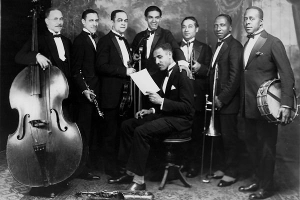 An image of Lou Hooper reading music with seven men surrounding him holding instruments