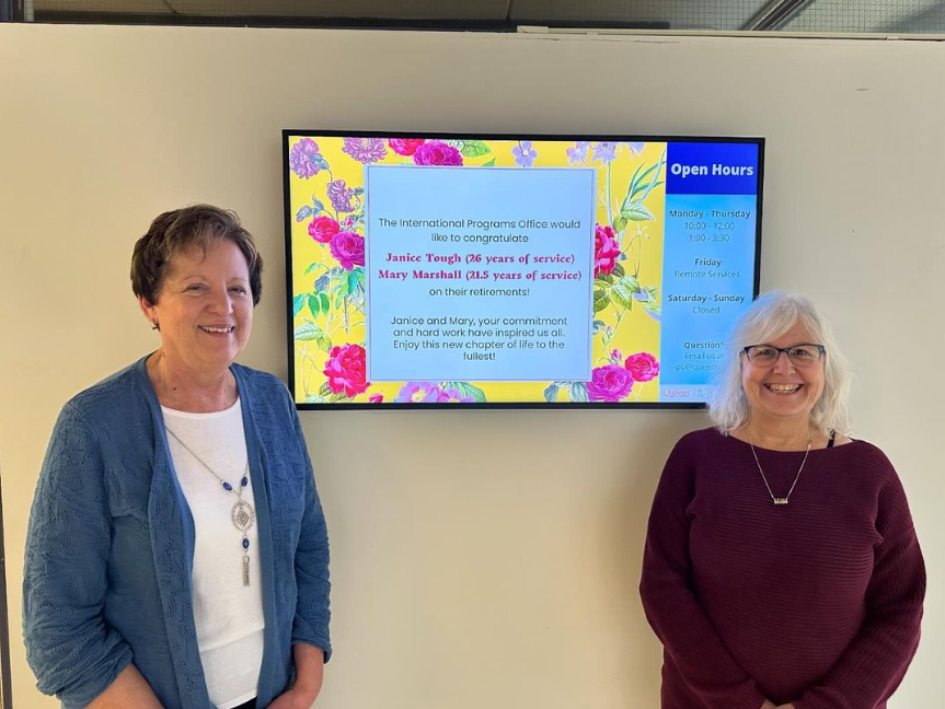 Photo of Mary and Janice standing in front of a TV announcing their retirement from the International Programs Office