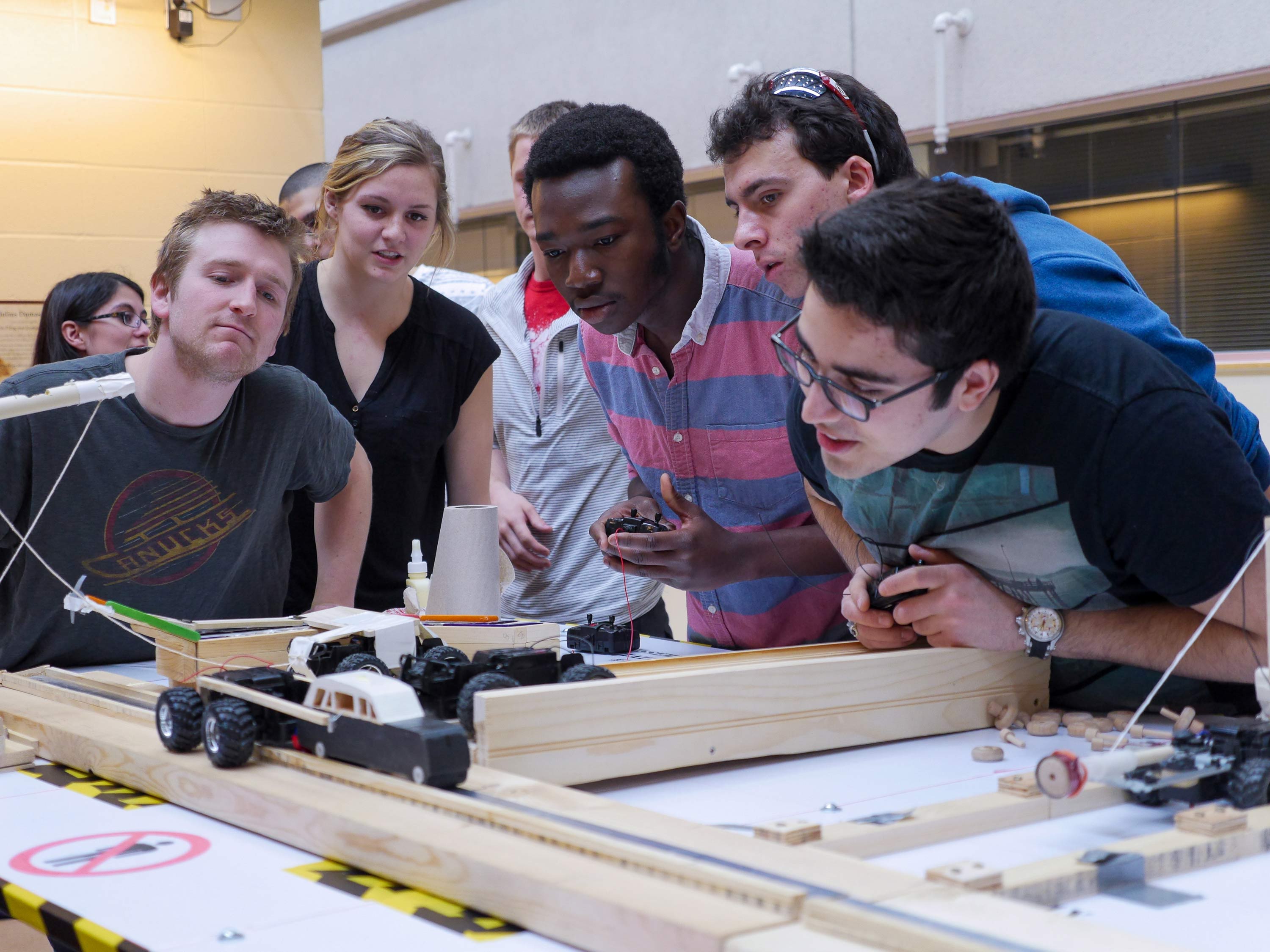 Group of six students closely watching remote controlled cars on track, two holding remotes