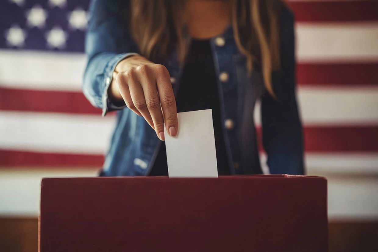 A person places a ballot in a box with an American flag in the background