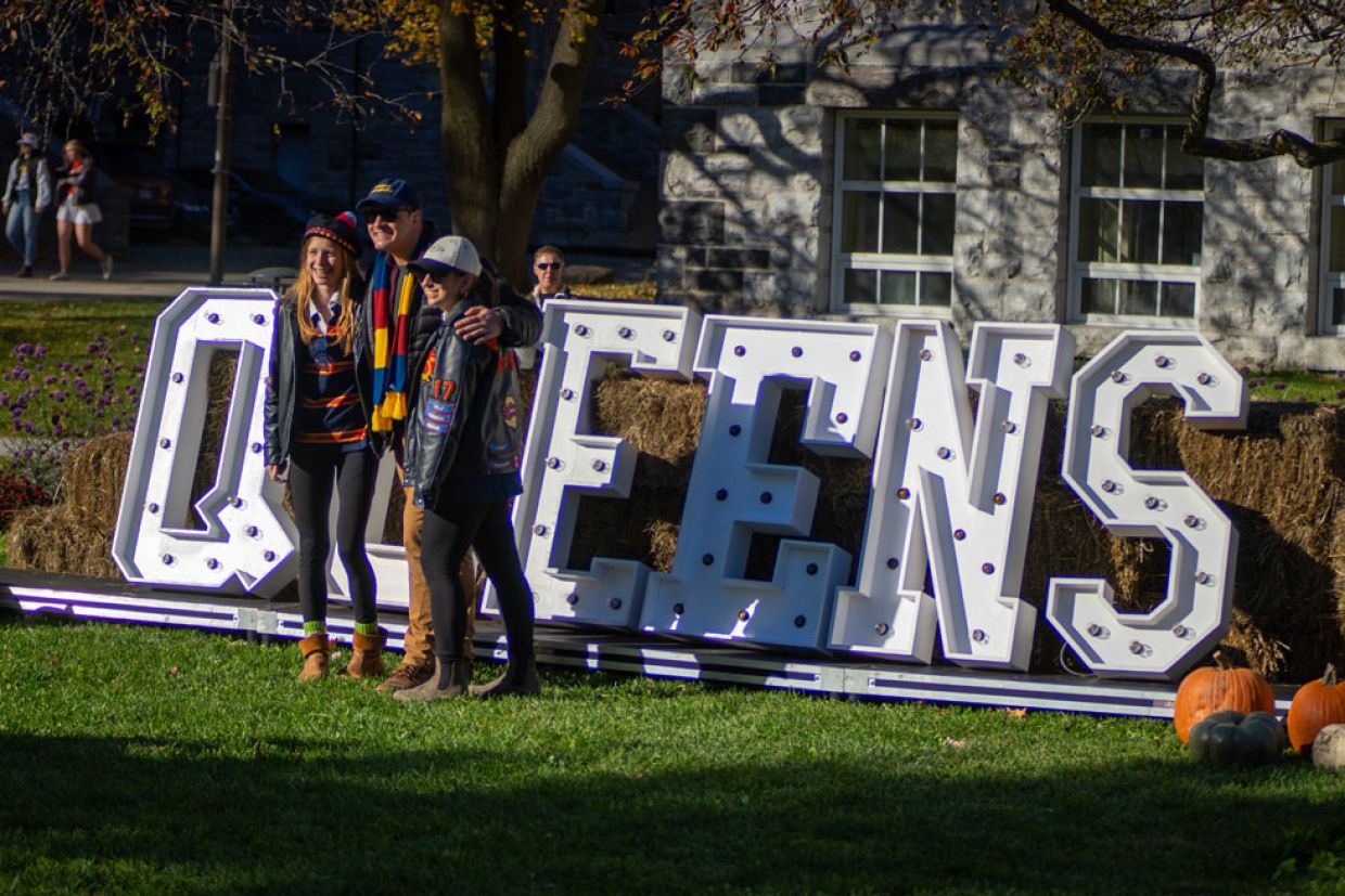 Three people pose for a photo in front of Queen's signage