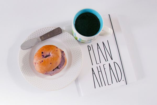 A sliced bagel on a plate with a spreading knife, coffee, and a journal titled "Plan Ahead."