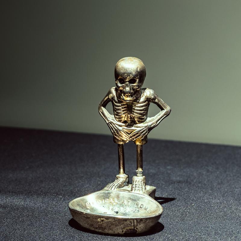 A metal ash tray with a skeleton standing to one stand. The ash tray has 'Medical at Home' engraved on it.