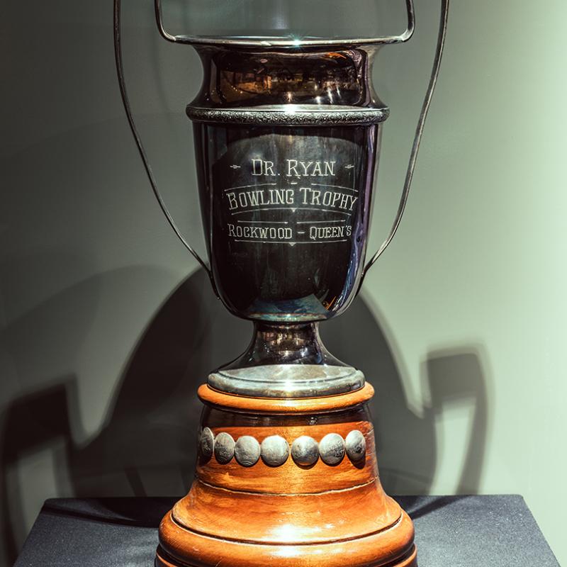 A bowling trophy sits atop a wood base. The trophy reads 'Dr. Ryan, Bowling Trophy, Rockwood – Queen's.'