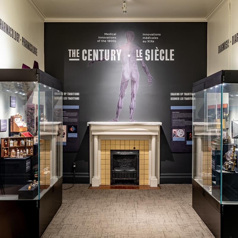 View of a room with two glass display cases to the left and right. The wall straight ahead has a fireplace on it and the back of a male body. The wall says 'Medical innovations of the 1800s, The Century.'