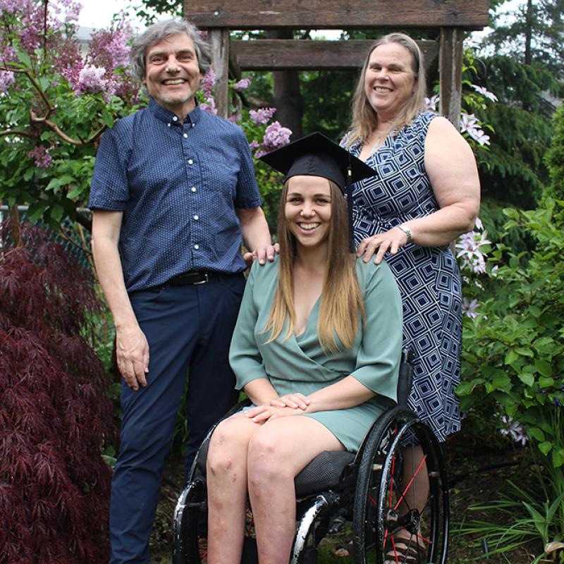 Katie Combaluzier sits in a wheelchair wearing a mortarboard, with her parents standing on either side of her.