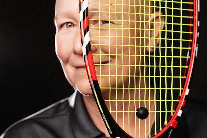 A woman wearing a Queen's jersey holds a squash racquet up to her face. She peers through the mesh of the racquet with her left eye.