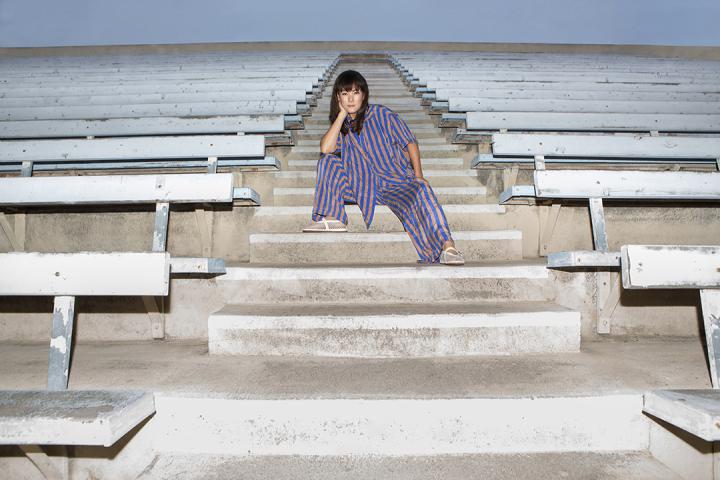 Laura Low Ah Kee sits on a staircase surrounded by bleachers. Her right arm rests on her knee with her head resting on her right hand. Her left left is outstretched in front of her with her left hand sitting on her knee.