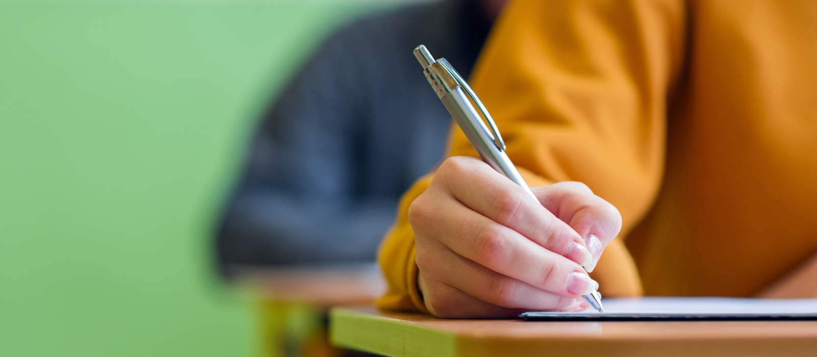 A hand of student writing an exam in the foreground with other students behind them also writing.