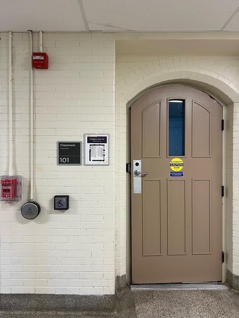 The entryway of room 101 in Kingston Hall is a beige door with a handle on the left side. An accessible automatic door button is located on the wall to the left of the door.