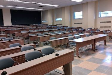 Tiered classroom with rows of brown desks and movable blue chairs. There are four windows along the wall.
