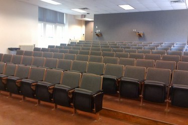 View from the front of the room: Rows of fixed chairs with tablet tables attached. The wall are grey.