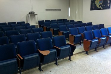View from the front of the room: Rows of fixed chairs with tablet tables attached. The wall are white.