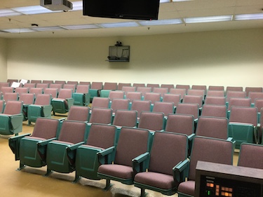 View from the front of the room: Rows of fixed chairs with tablet tables attached. The wall are cream.