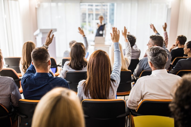 people with raised hands in rows facing a presenter at the front