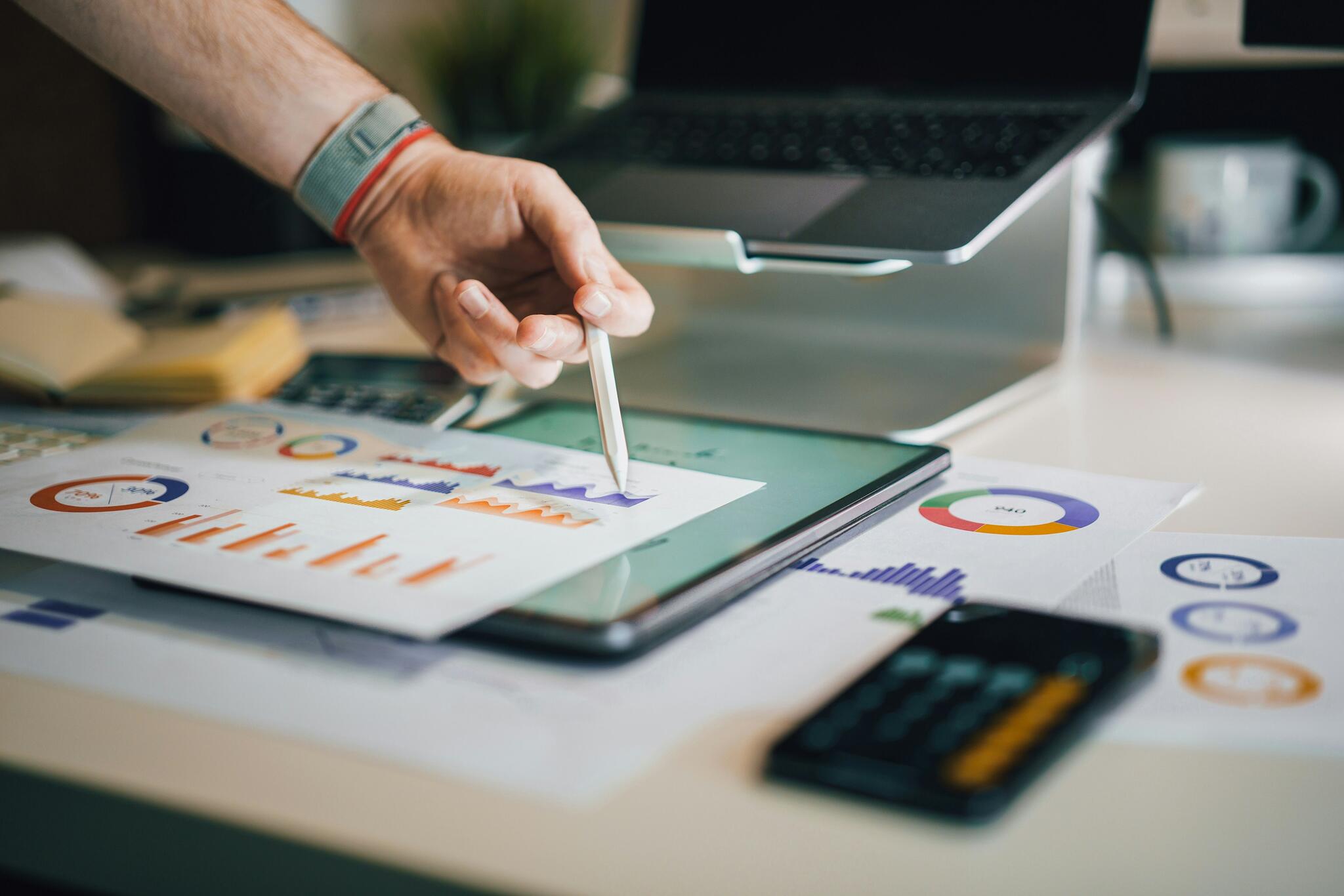 Person pointing to a tablet screen with papers scattered around it that display graphs in different colours