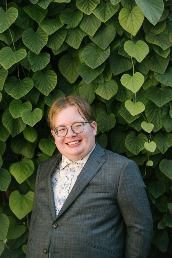 A photograph of Drumlin, a white nonbinary person with short brown hair and glasses. The photo is from the waist up, and they are wearing a grey suitjacket and a white dress shirt with blue flowers on it. The wall behind them is entirely covered in leaves. Photo by Viara Mileva. 