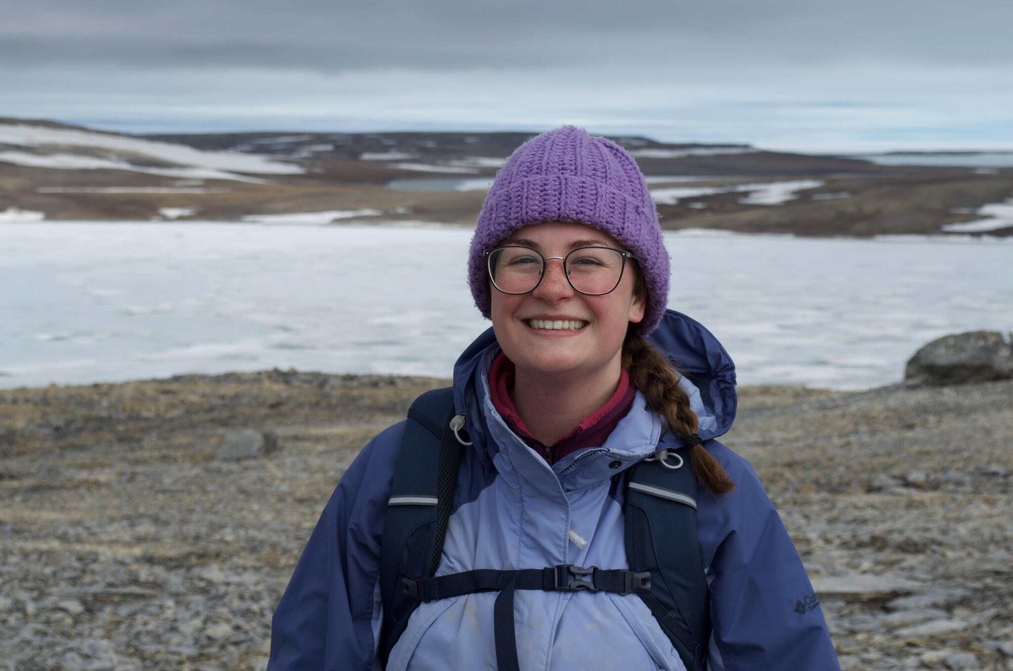 Alyssa Mckendrick wearing a bright purple toque, light blue jacket, and glasses stands in front of a snowy backdrop.