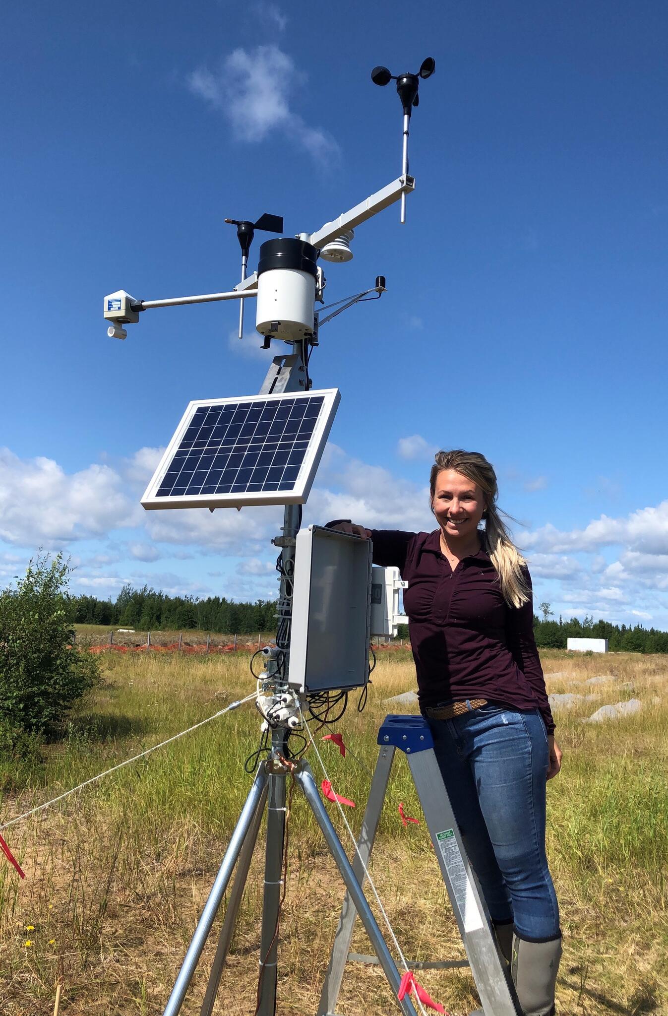 Caitlin Lapalme, wearing blue jeans and a maroon shirt, stands on a ladder next to a weather station in a grassy field with blue skies.