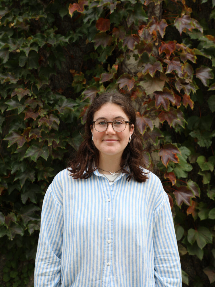 Sabien Edney, shoulder length brown hair and glasses stands in front of a wall of green and red leaves.