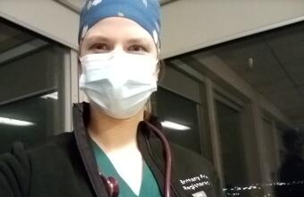 Woman with a mask and scrubs on, wearing a jacket overtop the scrubs.