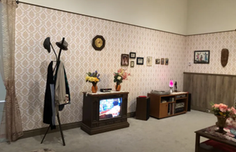 A photo showing a Mid-20th-century style family room with a hat rack, an analogue clock, a TV, and a motley of photographs, flower vases, and table decorations