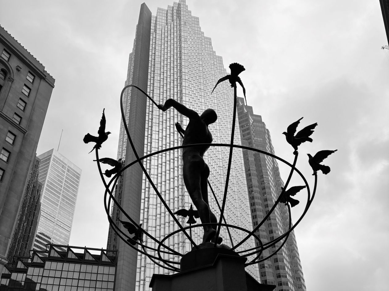 A black and white photograph of a statue in downtown Toronto featuring a man standing in the centre and doves flying around him