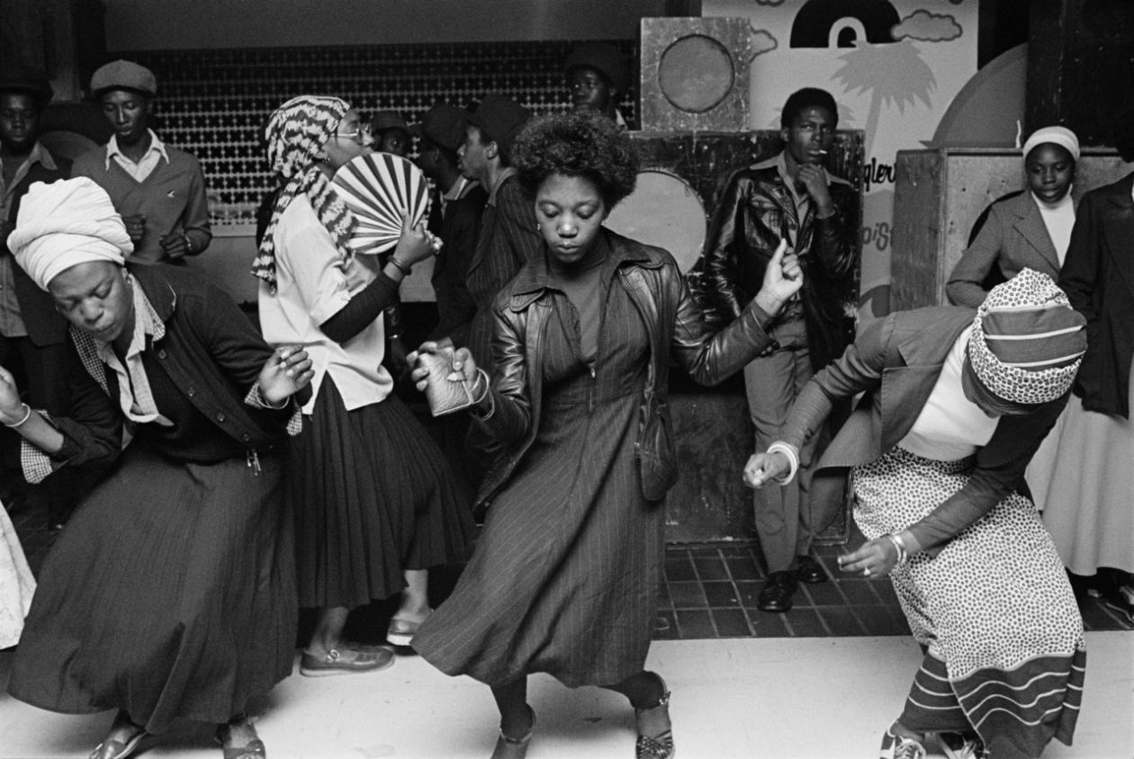 A black and white photograph of black women wearing skirts dancing with black men and women standing in the background