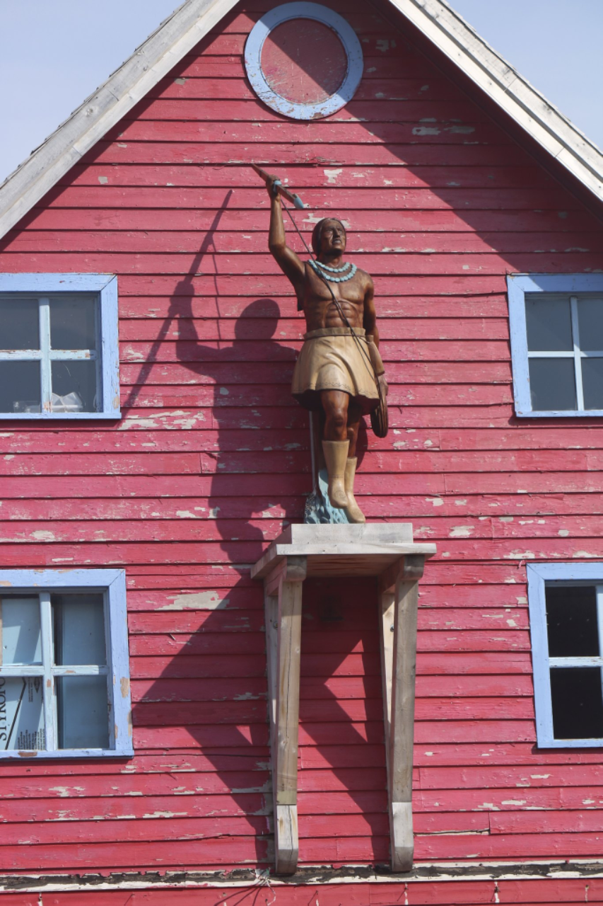 An image of an old red house with a statue of an Indigenous man standing atop a platform attached to the side