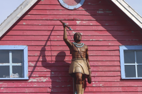 An image of an old red house with a statue of an Indigenous man standing atop a platform attached to the side